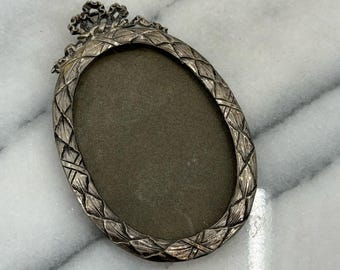 May include: Oval silver-toned picture frame with a decorative leaf and diamond pattern around the edge. The top of the frame features a small floral design. The frame has a dark gray interior.