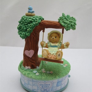 May include: A musical teddy bear figurine on a swing set. The bear is wearing a yellow dress with a blue bodice and a pink bow. The swing is attached to a brown tree with green leaves. A blue bird is perched on a nest at the top of the tree. The base of the figurine is decorated with a blue and white checkered pattern and the words "Cherished Teddies".