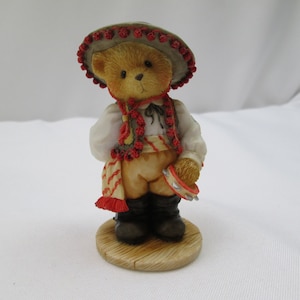 May include: A small, brown teddy bear figurine wearing a red and white sombrero and a red and white vest. The bear is holding a tambourine and is standing on a wooden base.