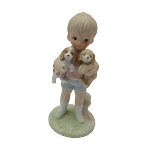 May include: A porcelain figurine of a boy holding two small dogs. The boy is wearing blue shorts and a pink shirt. The dogs are brown and white.