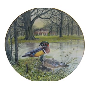 May include: A decorative porcelain plate featuring a painting of a pair of wood ducks in a woodland setting. The ducks are perched on the edge of a pond, with a house in the background. The plate is trimmed in gold and has the artist's signature "JERNER" in the lower right corner.