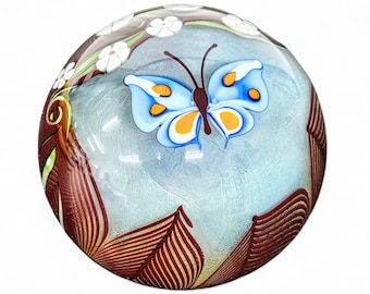 May include: A glass paperweight with a blue butterfly and white flowers on a brown and white background.