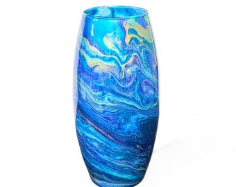 May include: A tall, oval-shaped vase with a vibrant, swirling design in shades of blue, teal, and gold. The glass vase has a glossy finish and a wide opening, suitable for displaying flowers or as a decorative piece.