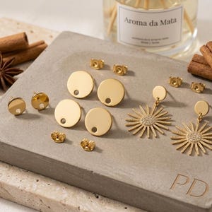 Pode incluir: Uma coleção de brincos dourados, incluindo brincos de pressão e pendentes, exibidos em uma superfície de concreto cinza. Pauzinhos de canela e anis estrelado adornam a cena. Ao fundo, uma garrafa com o texto "Aroma da Mata".