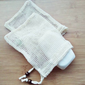 May include: Two off-white, mesh soap saver bags with a drawstring closure. One bag contains a white bar of soap. The bags are made of a woven material and have wooden beads on the drawstring.
