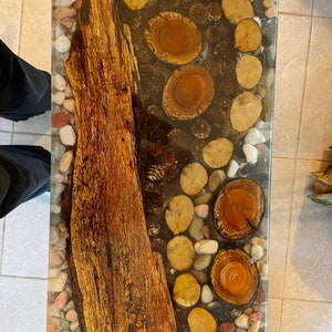 May include: A rectangular coffee table with a clear epoxy resin top. The resin contains a variety of natural elements, including wood slices, pine cones, and river rocks. The wood slices are arranged in a circular pattern, while the pine cones and river rocks are scattered throughout the resin.