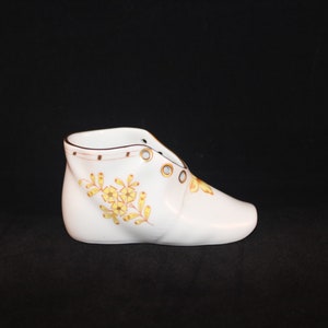 May include: A white ceramic shoe-shaped planter with yellow floral designs and brown trim. The planter has eyelets for laces and is set against a black background. A decorative item for a home.
