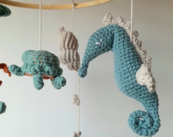 May include: A handmade baby mobile featuring crocheted sea creatures. The mobile includes a teal seahorse, a turtle, and a seashell, all suspended from a wooden hoop. The seahorse has a cream-colored mane and tail. The mobile is designed for a nursery.