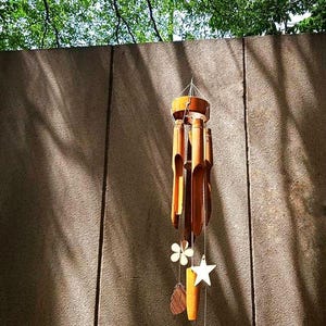 May include: A bamboo wind chime with a wooden top and decorative hanging elements. The chime features a star, flower, leaf, and feather-shaped accents. The background includes a textured wall and green foliage.