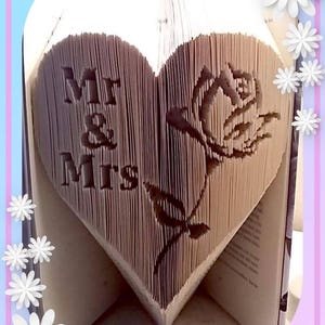 以下が含まれることがあります： ページに「Mr & Mrs」の文字が彫られた、ハート型の本の彫刻。右側にはバラが彫られています。本は開かれており、彫刻のデザインが明らかになっています。デザインは、白い花のアクセントが付いたピンクと青のボーダーで囲まれています。