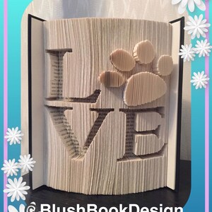 May include: A book sculpture with the word "LOVE" carved into the pages. A paw print design is cut out next to the letters. The book is open, revealing the text of the pages. The background is a light color.