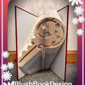 May include: A key-shaped book sculpture with a detailed design cut into the pages. The book is open, revealing the red edges of the pages and the intricate key silhouette. The text "BlushBookDesign" is at the bottom.
