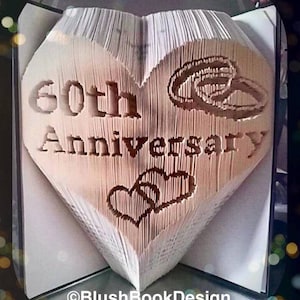 Puede incluir: Escultura de libro en forma de corazón con las palabras "60th Anniversary" talladas en las páginas. El diseño incluye anillos de boda tallados y dos corazones entrelazados. El libro está abierto, revelando las páginas cortadas.
