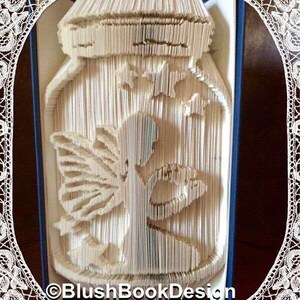 May include: A handmade paper art piece depicting a jar with a fairy and stars inside. The artwork is created by folding the pages of a book. The text "©BlushBookDesign" is visible at the bottom. The piece is framed with a white lace border.