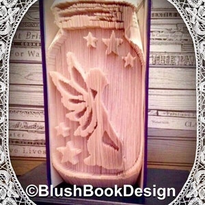 Könnte beinhalten: Eine Buchkunstskulptur einer Fee, die aus einem Einmachglas mit Sternen fliegt. Die Skulptur ist aus den Seiten eines Buches gefertigt und ist eine einzigartige und kreative Möglichkeit, ein Feenthema zu präsentieren.