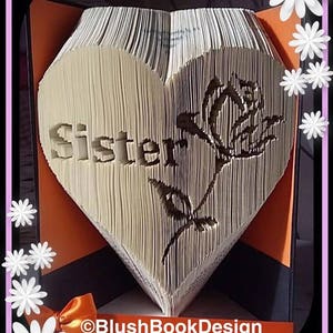 Può includere: Un libro piegato a forma di cuore con la parola "Sorella" e un disegno di rosa. Il libro è appoggiato su una superficie nera e arancione con un nastro legato attorno alla base. Il testo "BlushBookDesign" è visibile nella parte inferiore dell'immagine.