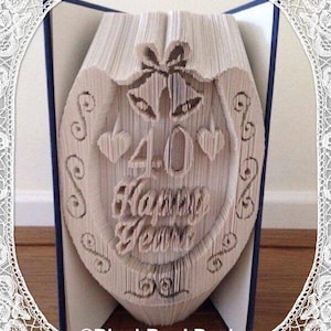 May include: A book sculpture with the words "40 Happy Years" carved into the pages. The design includes hearts, bells, and decorative swirls, ideal for an anniversary gift. The book is displayed open, with the carved design visible.