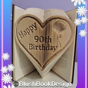 May include: A book folded into the shape of a heart with the words "Happy 90th Birthday" inscribed on it. The book is open and the pages are folded to create the heart shape. The book is on a table with a white background.