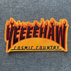 May include: A yellow and orange embroidered patch with the text "Yeehaw" in black lettering and "Cosmic Country" in smaller black lettering below. The patch is shaped like a flame.