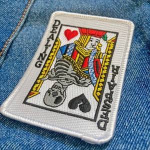 May include: Embroidered patch featuring a playing card design with a skeletal figure and the words "DEALING DEATH" in black. The card shows a queen with a red heart and a black spade. The patch has a white border.