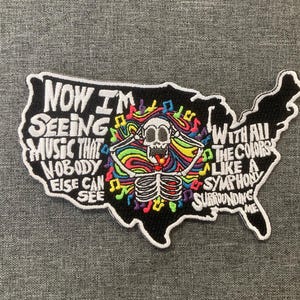 May include: Embroidered patch in the shape of the United States, featuring a skeleton with colorful hair and musical notes. The patch has the text "Now I'm Seeing Music That Nobody Else Can See" and "With All The Colors Like A Symphony Surrounding Me."