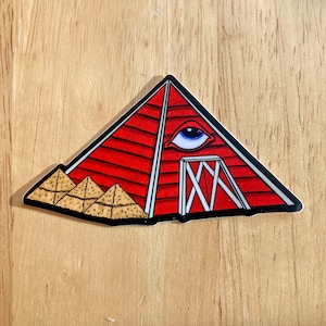 May include: Red barn shaped like a pyramid with a single blue eye on the front. The barn has a white wooden door with an X pattern. The barn is sitting on a pile of hay.