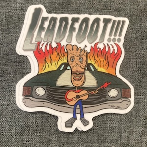 May include: A sticker featuring a cartoon foot playing a guitar in front of a car engulfed in flames. The sticker has the text "LEADFOOT!!!" in a bold, stylized font. The artwork is colorful with a humorous, edgy style.