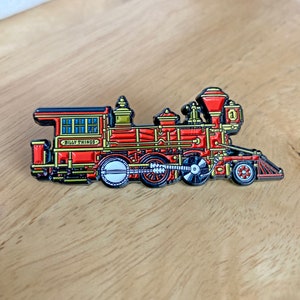 May include: A red and gold train engine with black wheels and a banjo for a smokestack. The engine has the words "Billy Things" on the side and the number "1" on the front.