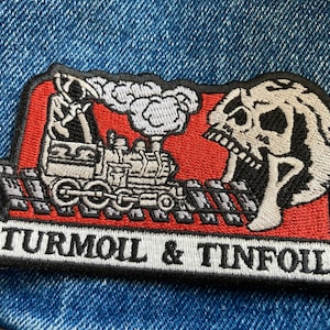 May include: An embroidered patch with a black and white train, a skull, and the text "TURMOIL & TINFOIL" in black on a red background.