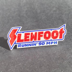 May include: A white, red and blue enamel pin with the text "SLEWFOOT RUNNIN' 90 MPH". The pin has a glittery finish.