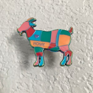 May include: A colorful enamel pin shaped like a goat with a patchwork design. The goat is standing on all four legs and has a small heart on its side. The words "HOME" are written on the goat's side.
