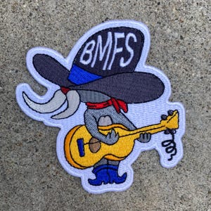 May include: An embroidered patch featuring a gray character wearing a large dark gray cowboy hat with the text BMFS in white, a red neckerchief, and blue boots, while playing a yellow acoustic guitar with a blue bridge and black strings.