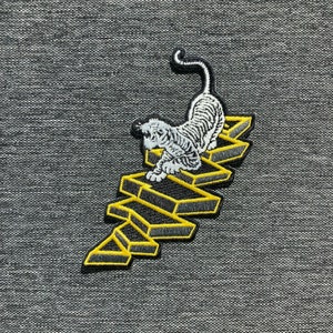 May include: Embroidered patch featuring a white tiger with black stripes, posed on a yellow and black geometric design. The patch has a detailed, textured appearance, suitable for application on clothing or accessories.