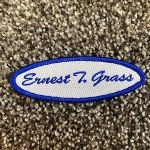 May include: White oval embroidered patch with a blue border. The text "Ernest T. Grass" is embroidered in blue on the patch.
