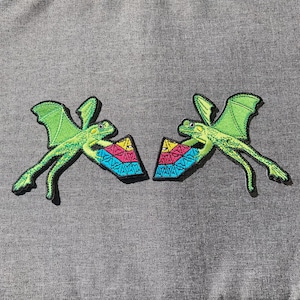 May include: Two embroidered patches featuring a green lizard with wings, holding a colorful geometric shape. The lizard has detailed stitching and is set against a gray background. The patches are likely for clothing or accessories.