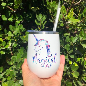 Puede incluir: Un vaso de acero inoxidable blanco con un unicornio y el texto "Magical AF" en letras rosa y azul. El vaso tiene una tapa transparente y una pajita.