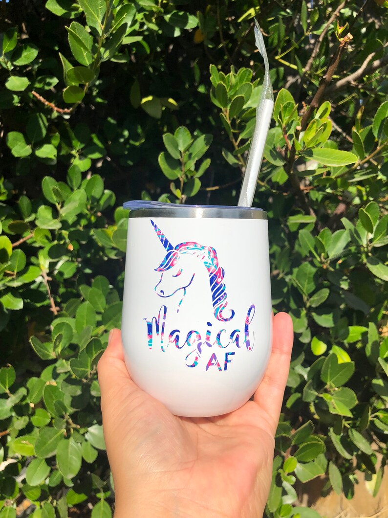 Puede incluir: Un vaso de acero inoxidable blanco con un unicornio y el texto "Magical AF" en letras rosa y azul. El vaso tiene una tapa transparente y una pajita.