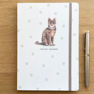 May include: A white notebook with a grey cat illustration and green polka dots. The text "Curious Thoughts" is printed below the cat. The notebook has a grey elastic closure.