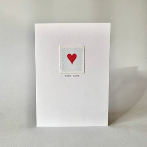 Tarjeta de aniversario pintada a mano con un corazón rojo. Tarjeta de acuarela con un corazón en relieve y la frase "Con amor". Tarjeta "Pensando en ti".