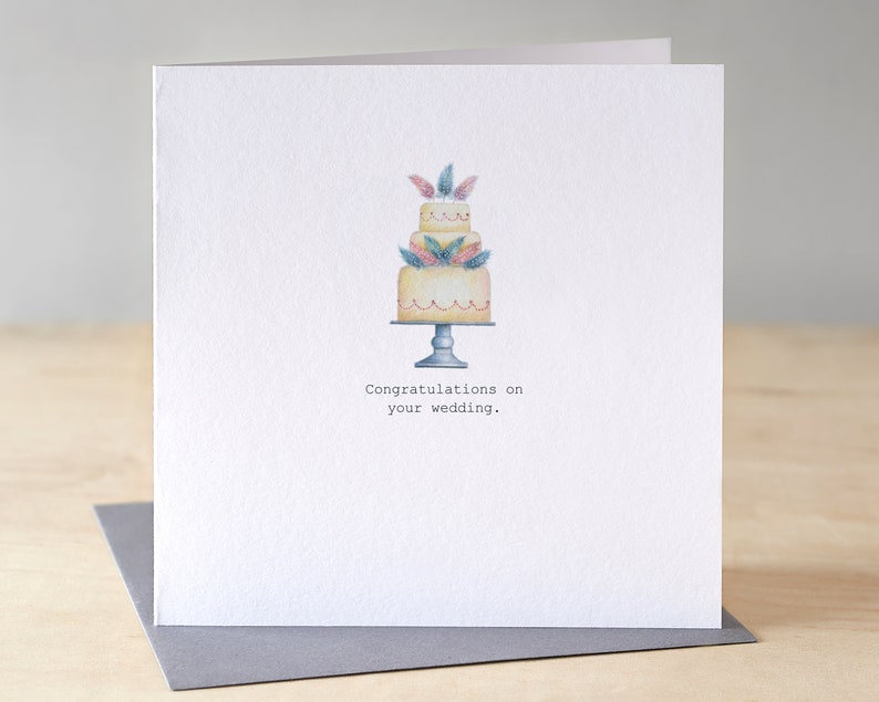 A Wedding Congratulations card with a hand painted 3 tiered cake on a stand. There are delicate little pink and blue feathers placed within each layer.