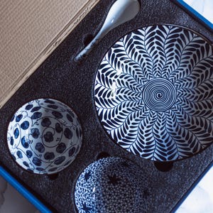 May include: A ceramic tableware set in a box, featuring a large plate, two small bowls, and a spoon. The plate has a blue and white leaf pattern, while the bowls have floral and star patterns. The spoon and bowls are white with blue accents.