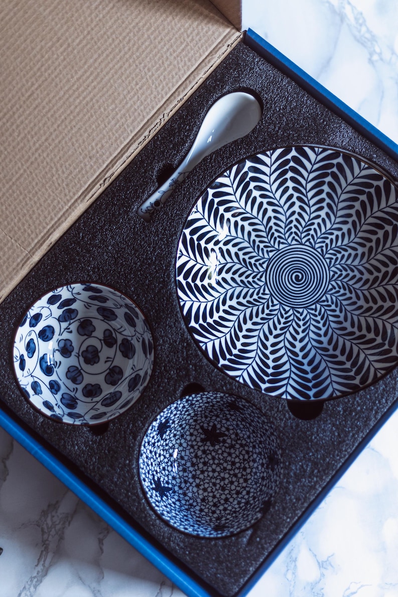 May include: A ceramic tableware set in a box, featuring a large plate, two small bowls, and a spoon. The plate has a blue and white leaf pattern, while the bowls have floral and star patterns. The spoon and bowls are white with blue accents.