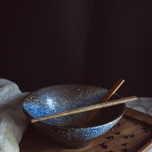 Pu&ograve; includere: Una ciotola in ceramica con esterno marrone scuro e finitura interna maculata blu e bianca. Un paio di bacchette di legno poggia orizzontalmente sopra la ciotola. La ciotola presenta un'ampia apertura circolare e una base affusolata.