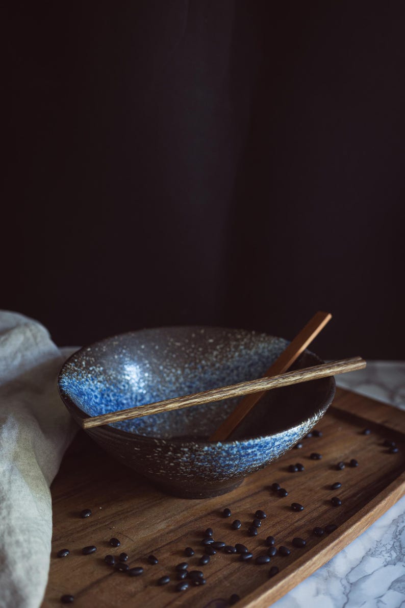Pu&ograve; includere: Una ciotola in ceramica con esterno marrone scuro e finitura interna maculata blu e bianca. Un paio di bacchette di legno poggia orizzontalmente sopra la ciotola. La ciotola presenta un'ampia apertura circolare e una base affusolata.