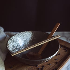 Pu&ograve; includere: Una ciotola in ceramica scura con bordo bianco maculato, dotata di un paio di bacchette in legno chiaro appoggiate sopra e un cucchiaio di legno all'interno. La ciotola &egrave; posizionata su un vassoio rettangolare in legno cosparso di fagioli neri.