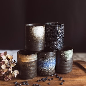 May include: Six handcrafted ceramic tumblers with textured surfaces and unique colour variations. The tumblers are dark brown, black, and blue, with speckled and glazed finishes. They are arranged on a wooden tray with dried flowers and small black beans.
