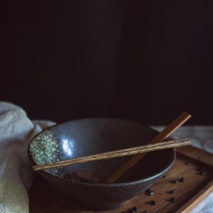 Pu&ograve; includere: Una ciotola in ceramica grigio scuro con una consistenza maculata, con un paio di bacchette di legno appoggiate sul bordo. La ciotola &egrave; posizionata su un vassoio di legno cosparso di piccoli fagioli neri, con un cucchiaio di legno all'interno.