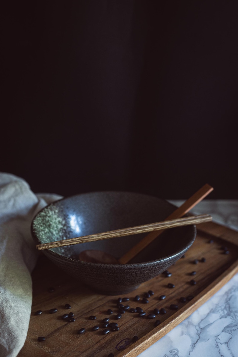 Pu&ograve; includere: Una ciotola in ceramica grigio scuro con una consistenza maculata, con un paio di bacchette di legno appoggiate sul bordo. La ciotola &egrave; posizionata su un vassoio di legno cosparso di piccoli fagioli neri, con un cucchiaio di legno all'interno.