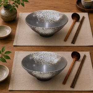 Pu&ograve; includere: Due ciotole in ceramica grigio scuro con un motivo a smalto bianco maculato all'interno e sul bordo. Ogni ciotola &egrave; accompagnata da un cucchiaio di legno e un paio di bacchette di legno, disposti su una tovaglietta in tessuto beige chiaro.