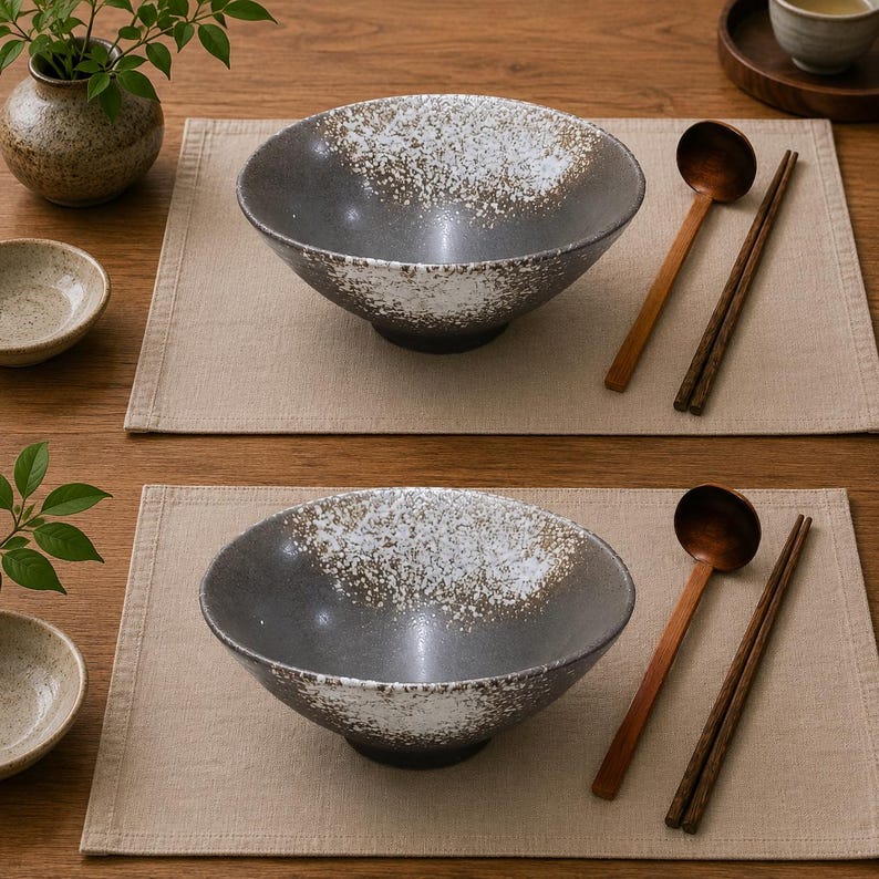 Pu&ograve; includere: Due ciotole in ceramica grigio scuro con un motivo a smalto bianco maculato all'interno e sul bordo. Ogni ciotola &egrave; accompagnata da un cucchiaio di legno e un paio di bacchette di legno, disposti su una tovaglietta in tessuto beige chiaro.